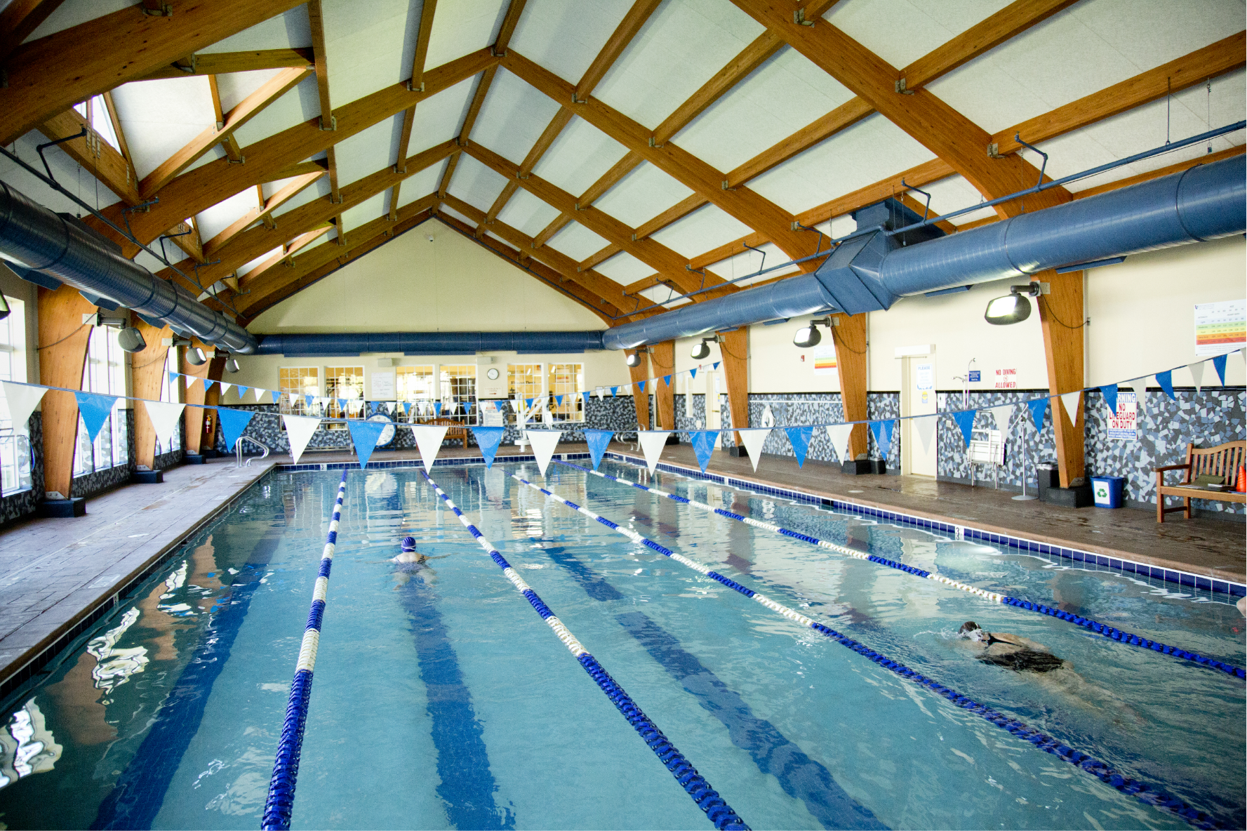 Indoor swimming pool with wooden beams on the ceiling, blue and white lane dividers, and a few swimmers in the water. The pool area is bright and spacious with large windows at the far end.