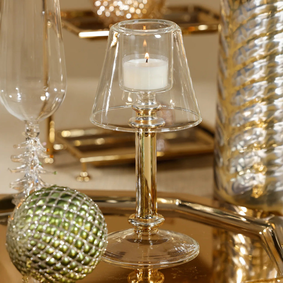 The ISOLA TEALIGHT LAMP by ZODAX features a clear glass lampshade, gold-tone cylindrical stem, and clear round base. Shown against a white background. Dimensions: 9.53 cm x 19.69 cm (3.75 in x 7.75 in).