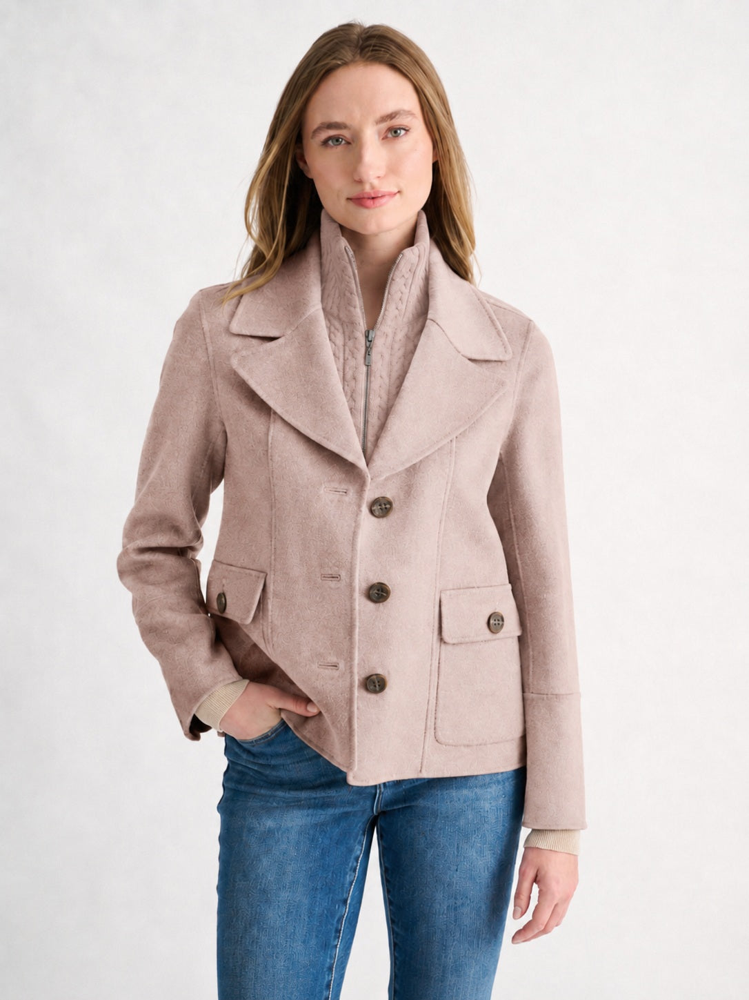 A woman wearing the KINROSS CASHMERE Knit Insert Notch Collar Coat in Sandstone over a collared shirt and blue jeans stands against a plain, light background, her hand in her pocket as she looks at the camera with a soft smile. The subtle cable knit insert on this classic KINROSS outerwear adds an elegant touch to her effortlessly chic look.