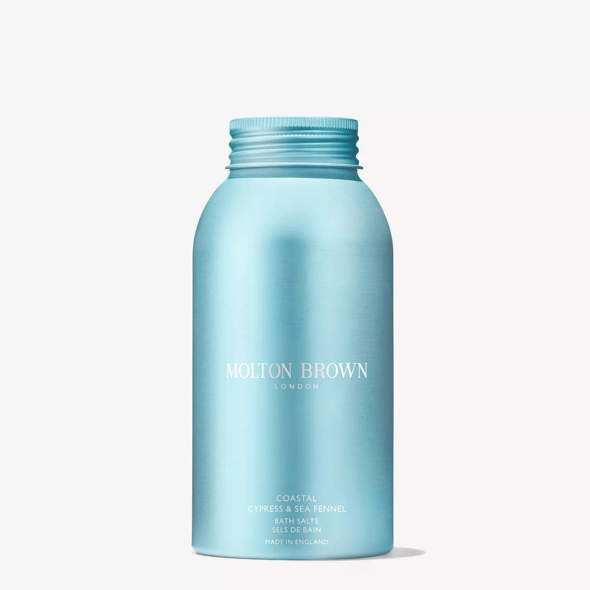A hand pours MOLTON BROWN - COASTAL CYPRESS BATH SALTS, known for their marine notes, from a blue bottle into a bathtub while water streams in from a chrome faucet.