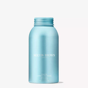 A hand pours MOLTON BROWN - COASTAL CYPRESS BATH SALTS, known for their marine notes, from a blue bottle into a bathtub while water streams in from a chrome faucet.