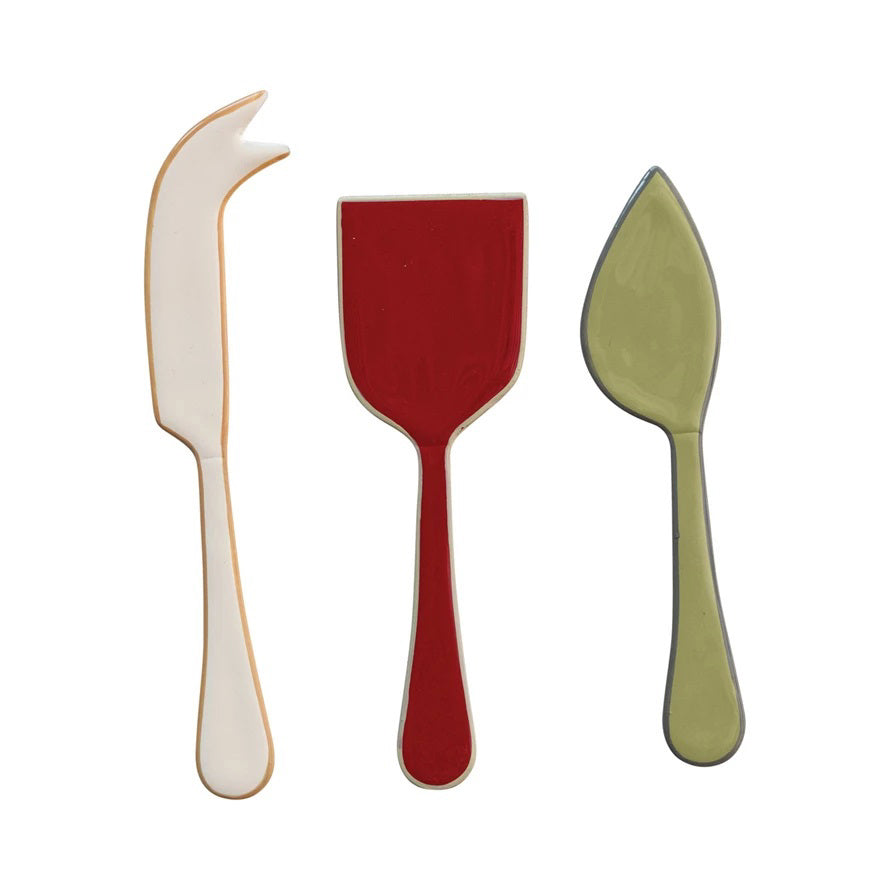 Three vibrant utensils—a cream cheese knife, green spreader, and red spatula from the CREATIVE COOP ENAMELED STAINLESS STEEL CHEESE SERVERS SET OF 3—rest side by side on a beige cloth over old paper atop a textured white surface.