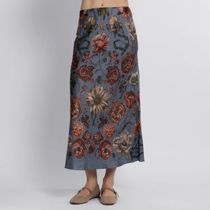 A person is seen from the back wearing a SABINA SAVAGE RING A RING O' ROSES SILK BIAS SKIRT, featuring English garden flowers in red, brown, and cream on gray-blue. They pair it elegantly with tan flat slip-on shoes.