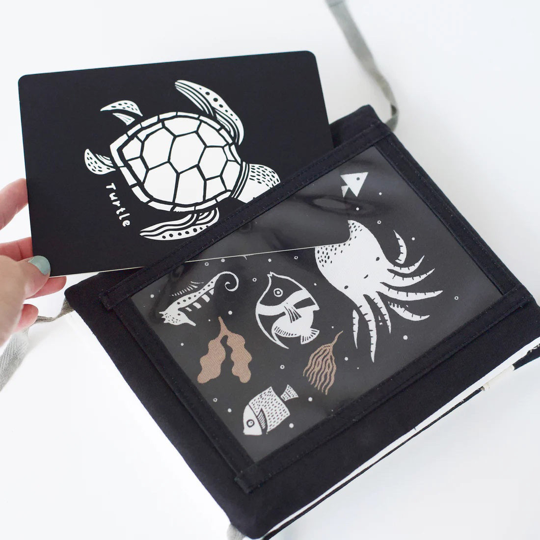 A TUMMY TIME GALLERY soft book from WEE GALLERY, featuring black and white fish and squid illustrations, is partially inserted in a clear front pocket. A brown tag attached by a string reads "tummy time gallery." This Montessori toy, ideal for sensory development, rests on a plain white surface.