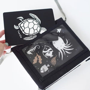 A TUMMY TIME GALLERY soft book from WEE GALLERY, featuring black and white fish and squid illustrations, is partially inserted in a clear front pocket. A brown tag attached by a string reads "tummy time gallery." This Montessori toy, ideal for sensory development, rests on a plain white surface.