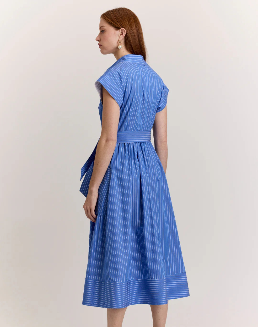 A woman with long brown hair models the HINSON WU Annabelle Short Sleeve Dress in Lake Blue Pencil Stripe, featuring a collared, button-down 100% cotton design with a matching waist tie. She stands against a plain light background.