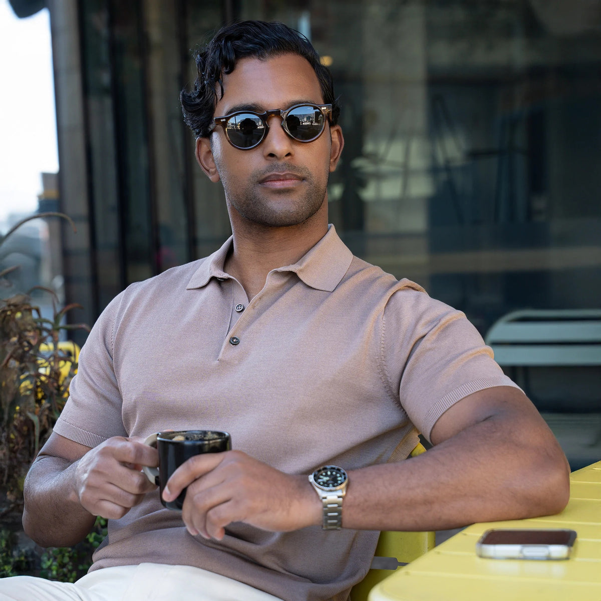 A man in sunglasses wears the COLLARS AND CO MARANELLO Mid-Weight Cotton & Silk 3-Button Polo in Taupe with white trousers, carrying a brown leather bag and phone. Green plants and modern buildings are in the background.