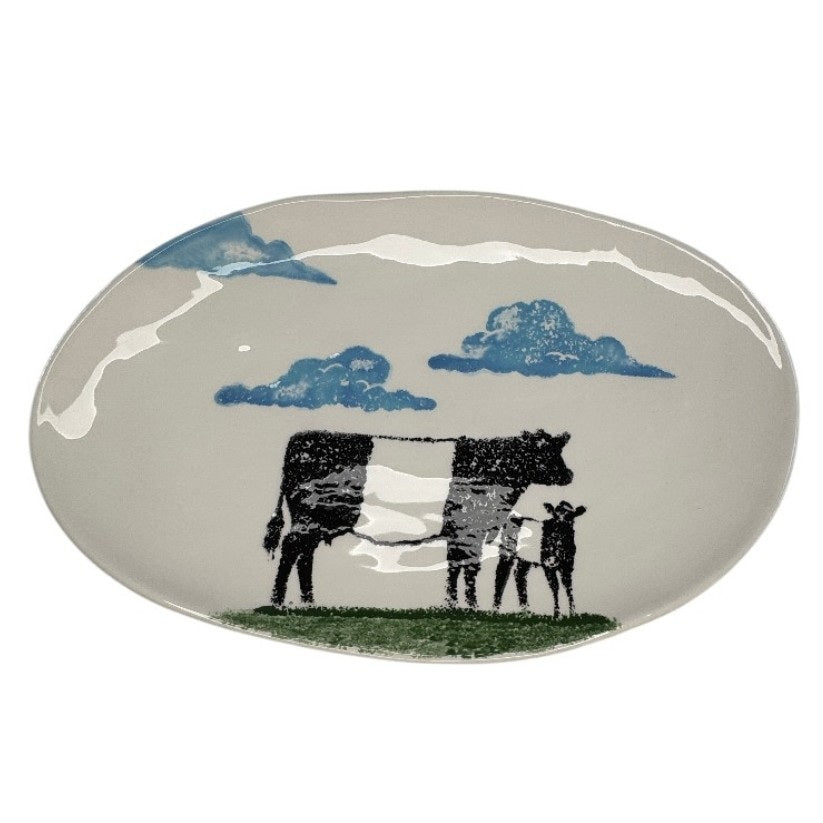 The FEARRINGTON SIGNATURE COLLECTION Beltie Oval Platter by VIETRI features black cow and calf silhouettes on green grass with blue clouds on a cream base, making it a charming gift inspired by Fearrington Village.