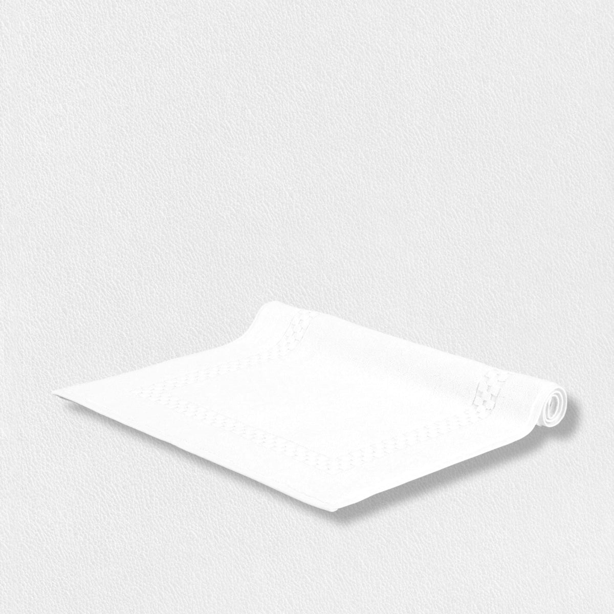 A small, white, folded paper napkin with decorative edges is placed on a light grey, textured surface. The napkin’s neatly rolled corner and plush cotton material evoke the luxurious feel of the Fearrington Lifestyle Towel Collection by Frette North America. The similar light tones create a clean and minimalist look.