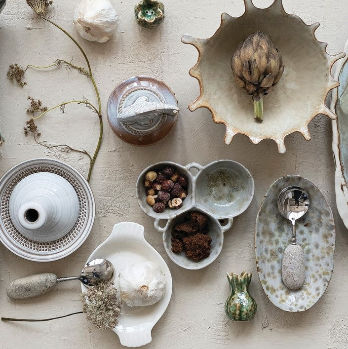 A rustic flat lay features the CREATIVE COOP 3-SECTION STONEWARE BOWL IN CREAM AND GREEN filled with spices, dried artichoke, garlic bulbs, vintage metal spoons, and assorted nuts on a textured beige surface.