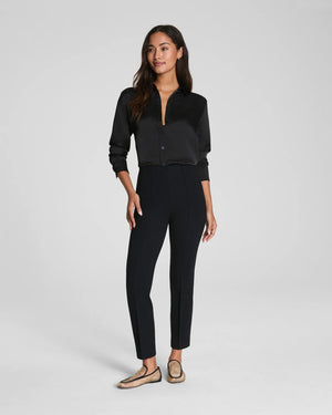 A person stands facing away in SPANX - PONTE ANKLE SLIM PANT IN BLACK by SPANX, paired with a black long-sleeve shirt and sheer pointed-toe flats. The look is simple and professional against a plain, light background.