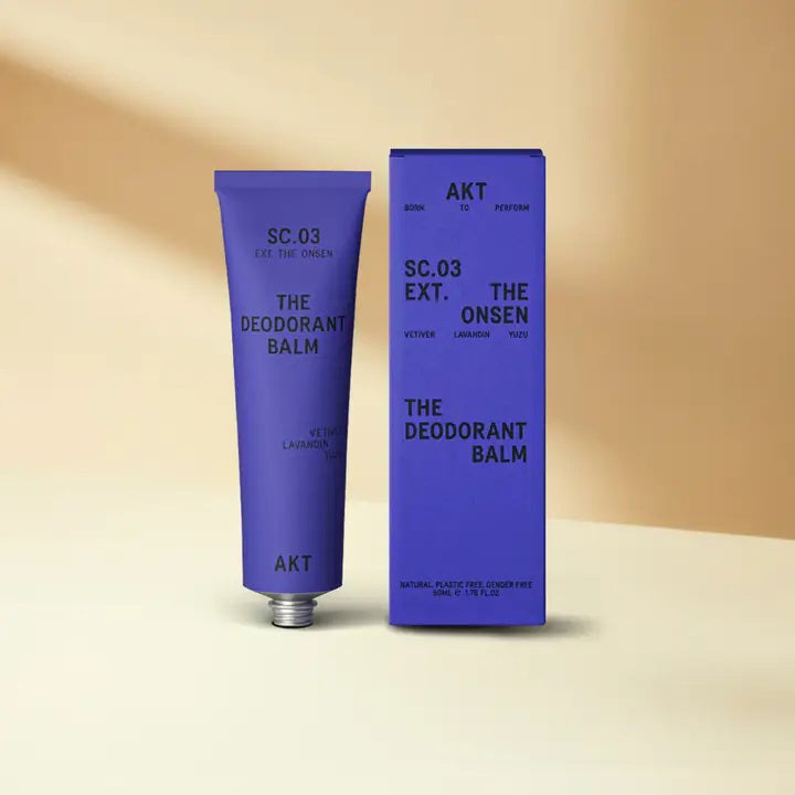 A purple tube of AKT LONDON’s AKT DEODORANT BALM THE ONSEN, a vegan and aluminum-free deodorant, stands by its box on a beige surface with matching background. Minimal packaging showcases black text and product details.