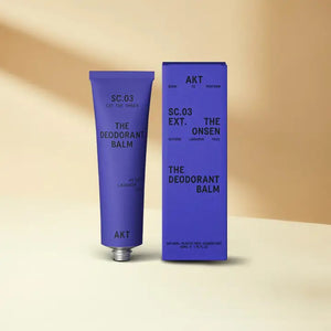 A purple tube of AKT LONDON’s AKT DEODORANT BALM THE ONSEN, a vegan and aluminum-free deodorant, stands by its box on a beige surface with matching background. Minimal packaging showcases black text and product details.
