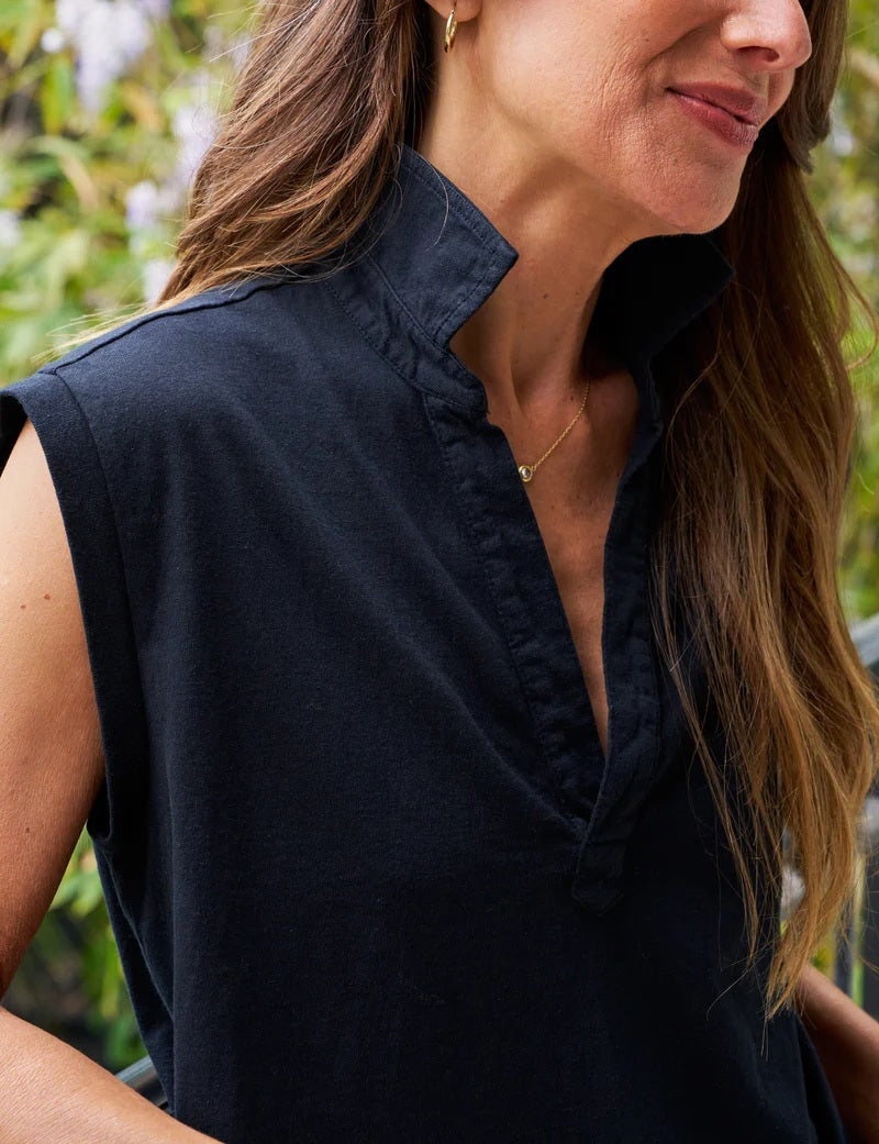 A woman with long brown hair stands on a balcony, smiling, wearing the FRANK & EILEEN Olivia Popover Henley Tank in British Royal Navy, with green foliage and purple flowers in the background.