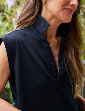 A woman with long brown hair stands on a balcony, smiling, wearing the FRANK & EILEEN Olivia Popover Henley Tank in British Royal Navy, with green foliage and purple flowers in the background.