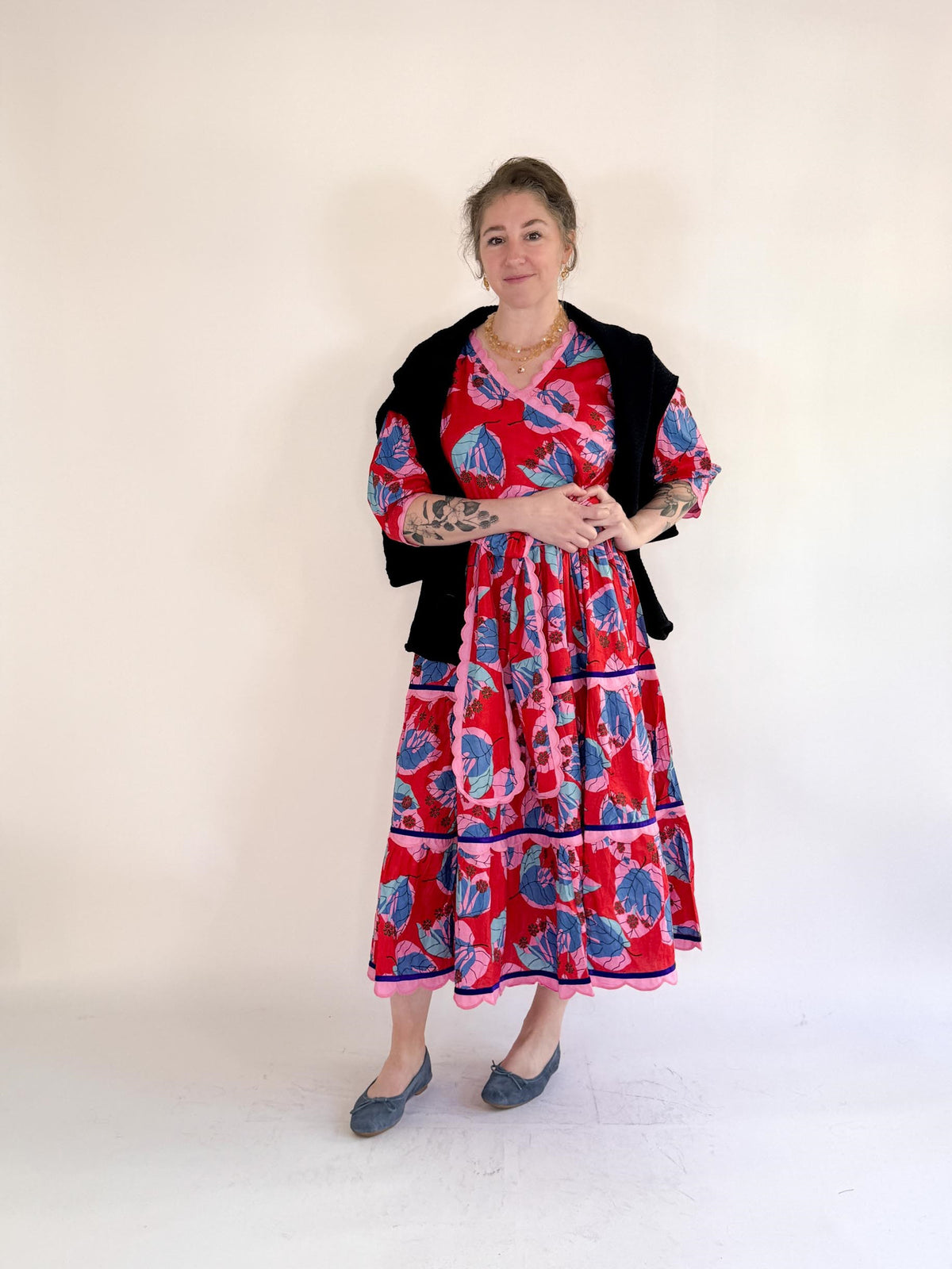 A smiling woman in the FARYLINA - CAMILLE LONG DRESS, a bold blue and pink floral cotton dress by FARYLINA, stands against a white background with one hand on her hip, showing arm tattoos and wearing gray flats.