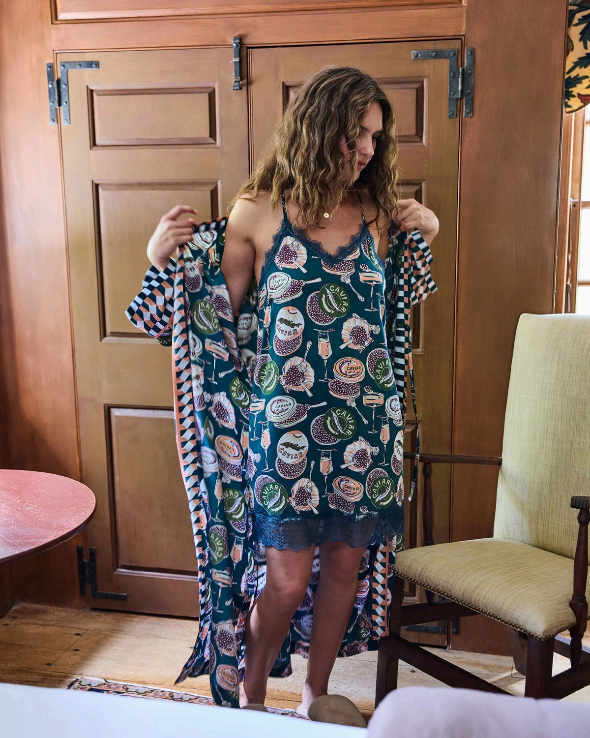 A woman stands barefoot against a white wall, smiling and holding a white cup in one hand as she poses slightly angled to the side. She is wearing the PRINTFRESH Champagne & Caviar Eco Satin Robe in Peacock Feather—a long, colorful eco-friendly robe featuring a vibrant botanical print with peacock feathers and checkered trim.