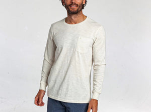 A man with short, curly hair and a beard wears the CRIQUET Cotton Slub Long Sleeve T-Shirt in Natural, styled with blue pants, standing against a plain white background.