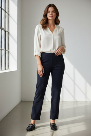 A woman stands indoors by a large window, wearing a long-sleeve cream blouse and the KRAZY LARRY, INC PULL ON WIDE ANKLE PANT in black, which features a tummy tuck design. She completes her look with black loafers and smiles slightly, her wavy brown hair catching the sunlight that casts shadows on the white floor and wall.
