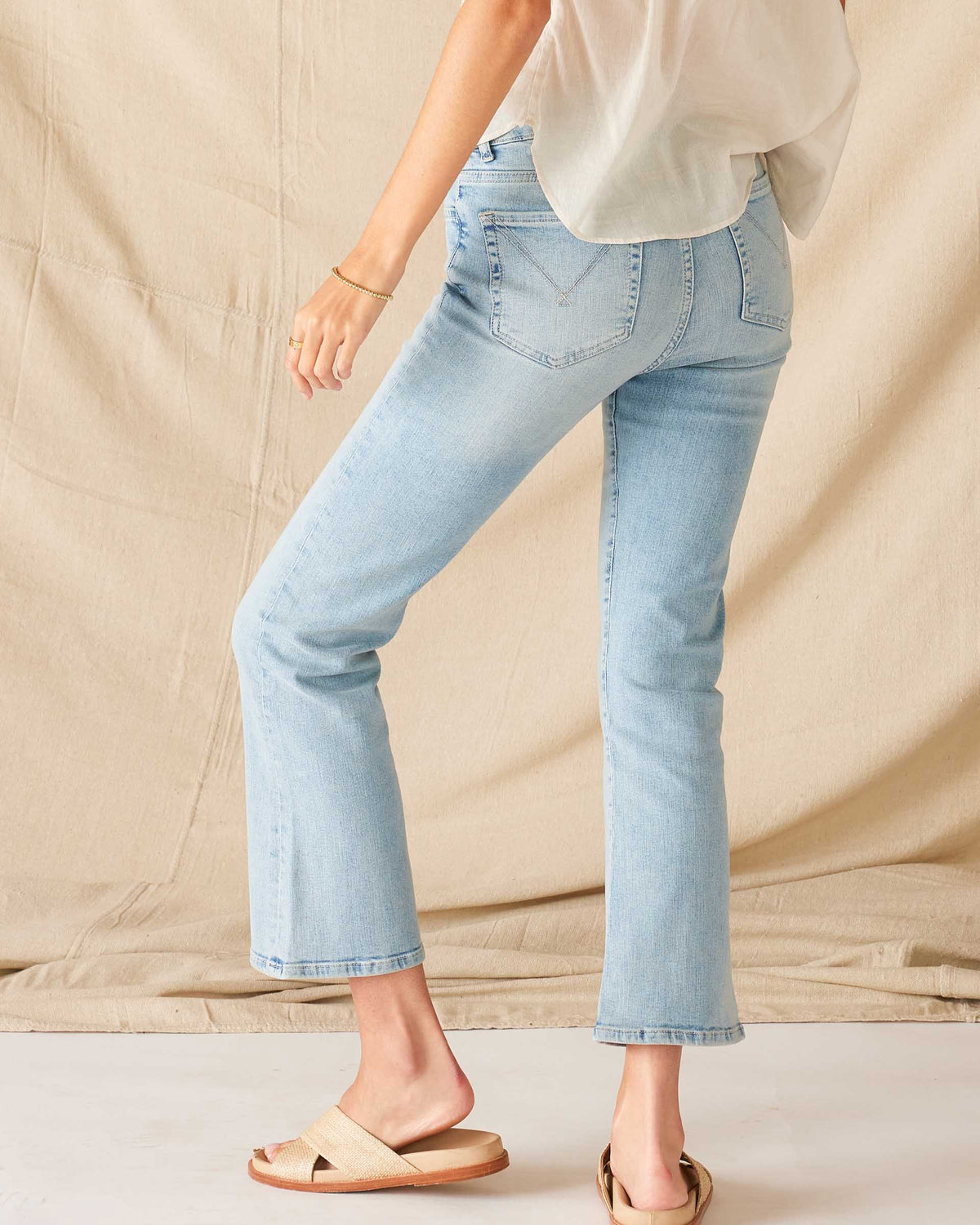 A person wears MERSEA’s Murphy Mini Boot Cut Jean in Sea Spray—light blue, 4-way stretch denim—paired with a white top and beige slide sandals, standing on a white floor before a beige fabric backdrop. Their face is not visible.