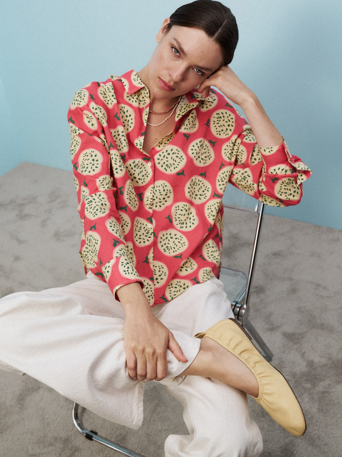 A woman stands against a light blue wall wearing the LUCY BLOUSE IN A FRUIT PRINT by 0039 ITALY, paired with white pants and beige shoes. The carpet is light gray, and she appears relaxed with arms at her sides.
