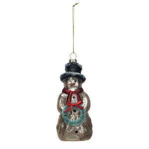 The CREATIVE COOP MERCURY GLASS SNOWMAN ORNAMENT, featuring a black hat, red scarf, and turquoise wreath, hangs on a textured white wall with a piece of rough-edged paper behind it.