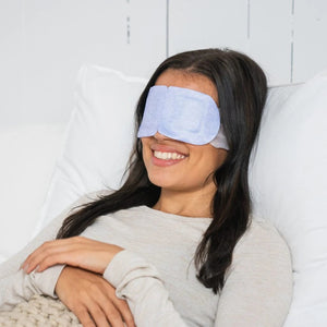 Hands hold a CORNEACARE - REST Self Heating Warm Compress as one hand removes a gray eye patch. The light blue pouch's minimalist design highlights its soothing eye dryness relief, enhanced by black and white elements on a plain backdrop.