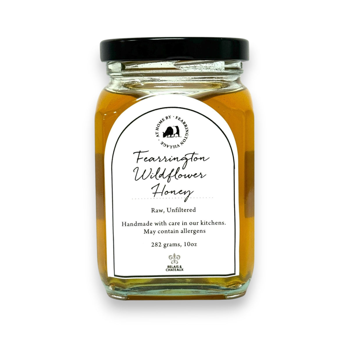 The Fearrington Signature Collection&#39;s hexagonal glass jar elegantly holds their Wildflower Honey, known for its raw quality. The label states: &quot;Raw, Unfiltered. Handmade with care in our kitchens. May contain allergens. 282 grams, 10 oz.&quot; Inside is the finest golden honey from Fearrington House.