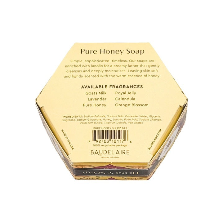 A luxurious BAUDELAIRE Pure Honey Soap 3.5 oz, featuring a hexagonal honeycomb pattern and beehive design, rests beside its floral BAUDELAIRE packaging. Nearby, a honey dipper releases golden honey over a textured backdrop. This exquisite soap is proudly made in the USA.