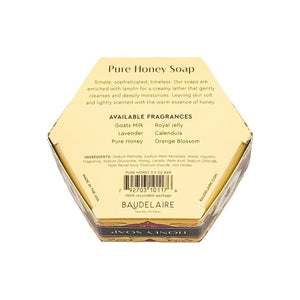 A luxurious BAUDELAIRE Pure Honey Soap 3.5 oz, featuring a hexagonal honeycomb pattern and beehive design, rests beside its floral BAUDELAIRE packaging. Nearby, a honey dipper releases golden honey over a textured backdrop. This exquisite soap is proudly made in the USA.
