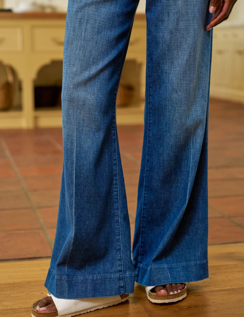 Indoors, someone wears a white long-sleeve shirt, white sandals, and FRANK &amp; EILEEN's WEST VILLAGE NYC WIDE LEG DENIM TROUSER IN 1984 WASH, with their left hand in their pocket. The kitchen background features baskets and wooden cabinets.