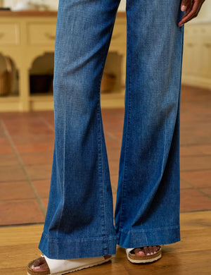 Indoors, someone wears a white long-sleeve shirt, white sandals, and FRANK & EILEEN's WEST VILLAGE NYC WIDE LEG DENIM TROUSER IN 1984 WASH, with their left hand in their pocket. The kitchen background features baskets and wooden cabinets.