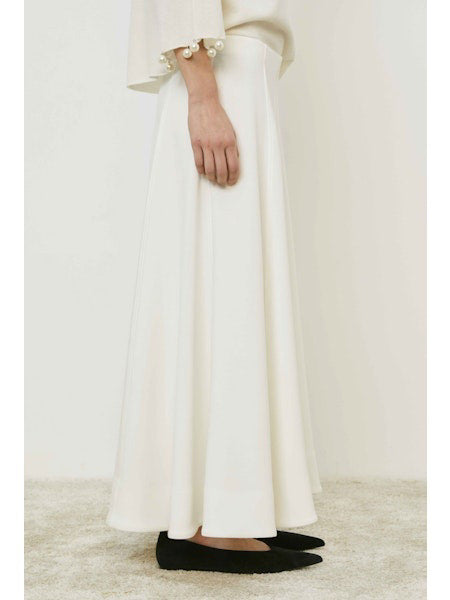 A person wears the MEIMEIJ - SKIRT, a long, flowing minimalist piece by MEIMEIJ, styled with a white top with pearl-detailed sleeves and black flats, standing on a light carpet.