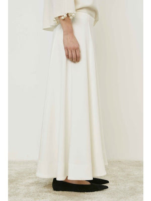 A person wears the MEIMEIJ - SKIRT, a long, flowing minimalist piece by MEIMEIJ, styled with a white top with pearl-detailed sleeves and black flats, standing on a light carpet.