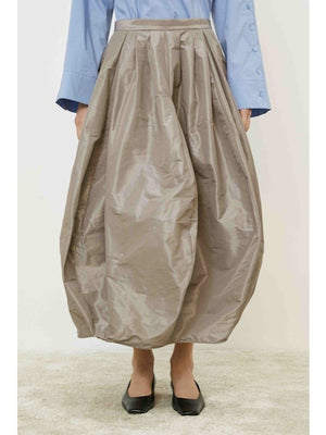 A person wears the MEIMEIJ Taffeta Balloon Skirt in shiny taupe, paired with a light blue long-sleeve shirt and black flats, standing on a light carpet against a white background.