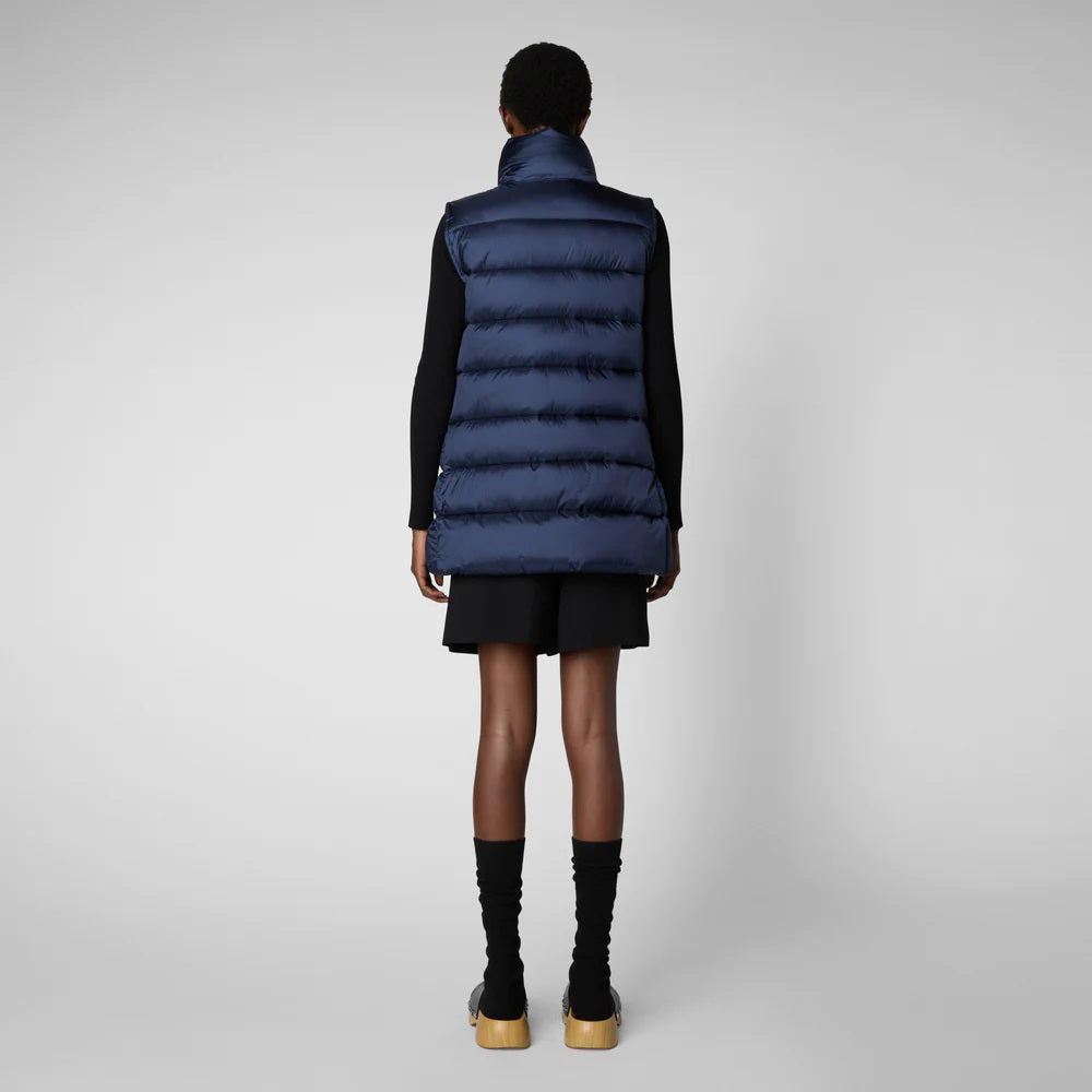 A person wearing a black cap, sunglasses, the SAVE THE DUCK - CORAL PUFFER VEST with animal-free down from SAVE THE DUCK over a black long-sleeve top, beige wide-legged pants, and black ankle boots stands against a plain background. The outfit appears to be styled for colder weather.