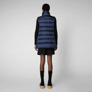 A person wearing a black cap, sunglasses, the SAVE THE DUCK - CORAL PUFFER VEST with animal-free down from SAVE THE DUCK over a black long-sleeve top, beige wide-legged pants, and black ankle boots stands against a plain background. The outfit appears to be styled for colder weather.