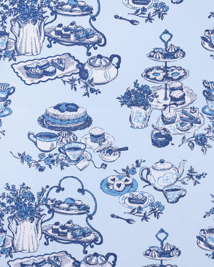 A woman stands barefoot on a wood floor, wearing the PRINTFRESH High Tea Toile Pintuck Nightgown in Notting Hill Blue—a vintage-inspired piece with short sleeves, a ruffled hem, and a charming blue illustrated print. She poses against a white wall with a relaxed smile.