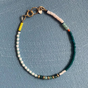 The ALICE RISE - EMERALD BRACELET by ALICE RISE showcases handmade North Carolina artistry with white, yellow, green, teal, and metallic gold beads and a gold clasp, elegantly displayed on a textured blue surface.