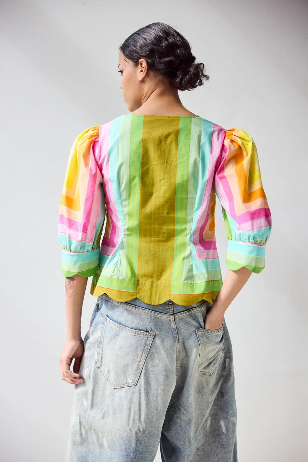 A person wears the HOPE FOR FLOWERS - SCALLOPED SHIRT IN CONCENTRIC SQUARES, featuring colorful geometric stripes and scalloped edges, paired with light blue jeans. The image highlights the torso.