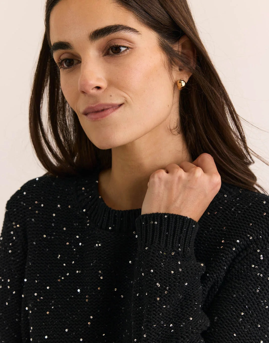A woman with long dark hair, dressed in the HINSON WU - QUEENIE LONG SLEEVE SEQUIN SWEATER IN BLACK by HINSON WU and blue jeans, stands against a plain light background, smiling gently at the camera.