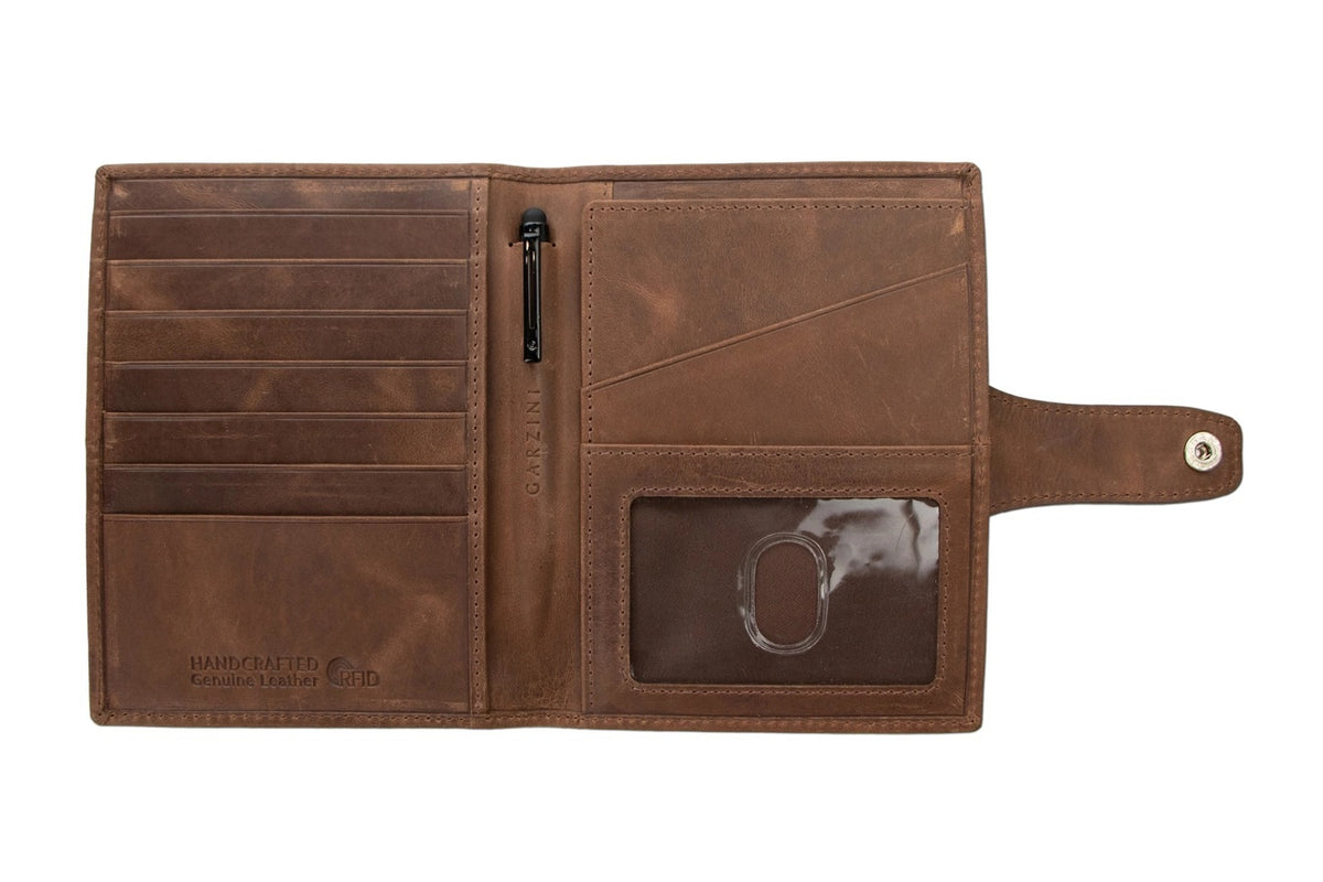 GARZINI RFID-blocking leather passport holder with AirTag compartment in brown, featuring a strap and snap button closure on a white background. The holder has a slightly worn, vintage look.