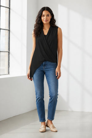 A woman with long dark hair stands indoors near a sunlit window, facing the camera and smiling slightly. She is wearing the SEVENTY Jersey Sleeveless Top with Draped Hemline by SEVENTY—a white wrap top featuring a V-shaped shawl collar and an asymmetric crossed design—paired with blue jeans and beige loafers.