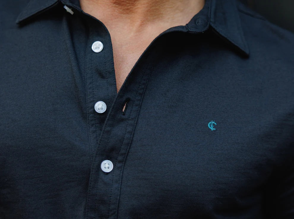 A man with short brown hair, wearing sunglasses, blue jeans, and the CRIQUET BRRR° Long Sleeve Range Polo in Dark Knight by CRIQUET, holds a takeaway coffee cup while standing against a white background.