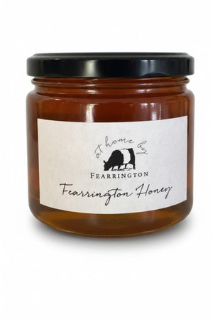 The Fearrington Signature Collection's hexagonal glass jar elegantly holds their Wildflower Honey, known for its raw quality. The label states: "Raw, Unfiltered. Handmade with care in our kitchens. May contain allergens. 282 grams, 10 oz." Inside is the finest golden honey from Fearrington House.