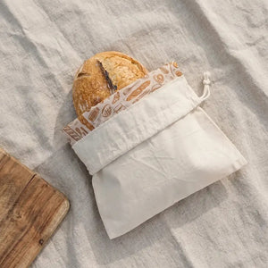 A round loaf of bread is partially tucked inside the BEES WRAP - BEESWAX REUSABLE BREAD BAKERY BAG NATURAL by BEES WRAP, a beige cloth bag with a drawstring and white label on the front, set against a white background—perfect for sustainable food storage.