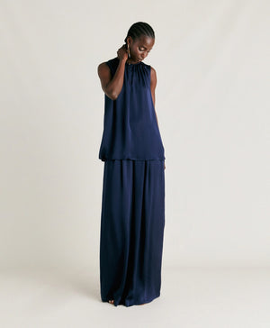 A person stands against a plain background wearing the MOMONI - CRIS SLEEVELESS TOP, a loose-fitting, round neck top in an elegant navy blue. They are also dressed in matching wide-leg pants crafted from pure silk, creating a sophisticated and flowing look. The individual has short, styled hair and is glancing downward with one hand touching their ear.
