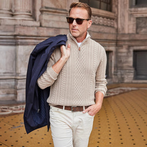 A man smiles while leaning on a wooden bench outdoors, wearing COLLARS AND CO's Napoli Cable Knit Quarter Zip Wool and Cashmere Sweater in Oatmeal.