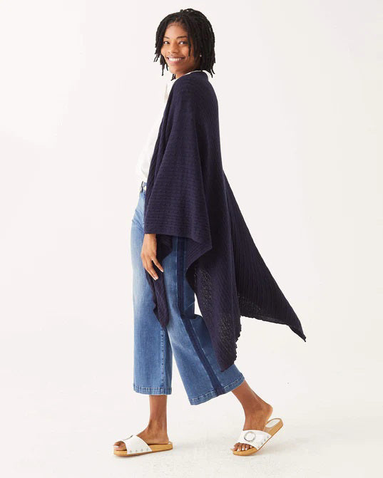 A woman with short, curly hair smiles while wearing a white blouse, wide-leg blue jeans, and the MERSEA CHARLESTON CABLE WRAP over her outfit, standing against a plain white background.