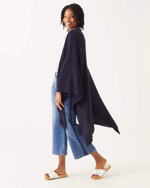 A woman with short, curly hair smiles while wearing a white blouse, wide-leg blue jeans, and the MERSEA CHARLESTON CABLE WRAP over her outfit, standing against a plain white background.